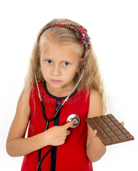 Bastante pequeña niña con estetoscopio en la barra de chocolate buscando preocupado en el hábito de nutrición poco saludable —  Fotos de Stock