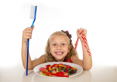 cute female child eating dish full of sweets and holding huge toothbrush in dental care and health concept