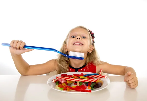 Cute female child eating dish full of sweets and holding huge toothbrush in dental care and health concept — 图库照片