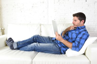 young attractive 30s man using digital tablet pad lying on couch at home networking looking relaxed