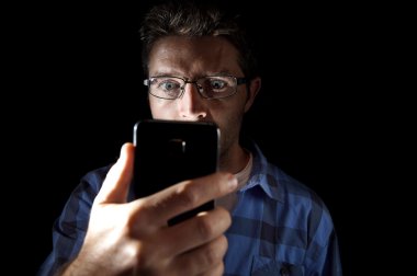 close up portrait of young man looking intensively to mobile phone screen with blue eyes wide open isolated on black background clipart