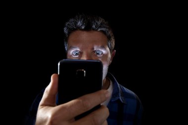 close up portrait of young man looking intensively to mobile phone screen with blue eyes wide open isolated on black background