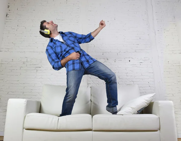 20s or 30s man jumped on couch listening to music on mobile phone with headphones playing air guitar — Stock fotografie