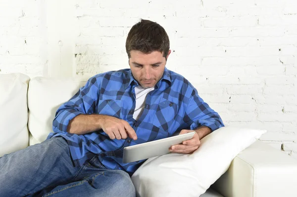 Young attractive 30s man using digital tablet pad lying on couch at home networking looking relaxed — Stock fotografie