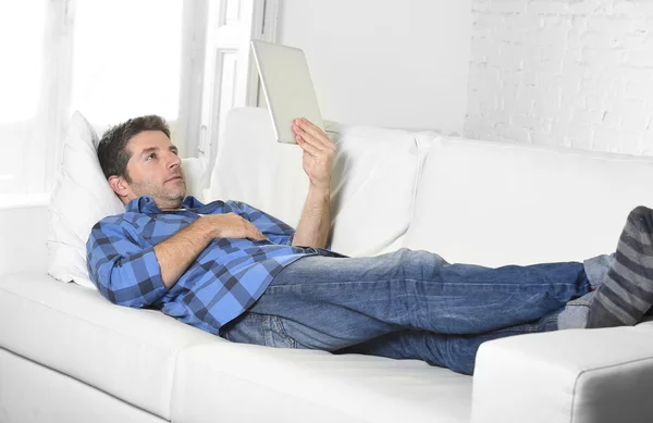 Young attractive 30s man using digital tablet pad lying on couch at home networking looking relaxed — Stock fotografie