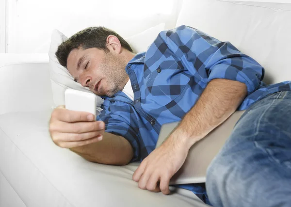 Atractivo hombre durmiendo en el sofá de casa con teléfono móvil y tableta digital en sus manos — Foto de Stock