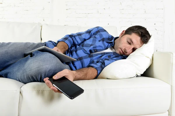 Atractivo hombre durmiendo en el sofá de casa con teléfono móvil y tableta digital en sus manos — Foto de Stock