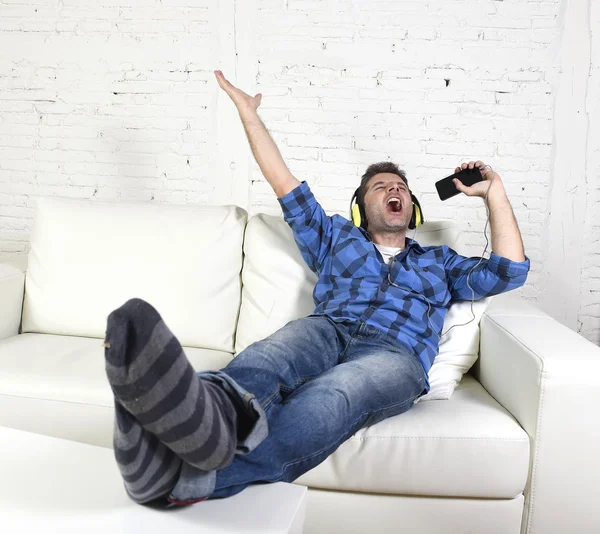 Happy crazy man on couch listening to music holding mobile phone as microphone — Stock fotografie