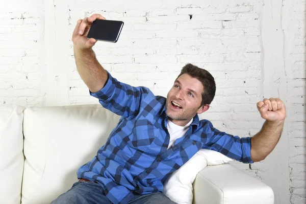 Young attractive 30s man taking selfie picture or self video with mobile phone at home sitting on couch smiling happy — Zdjęcie stockowe