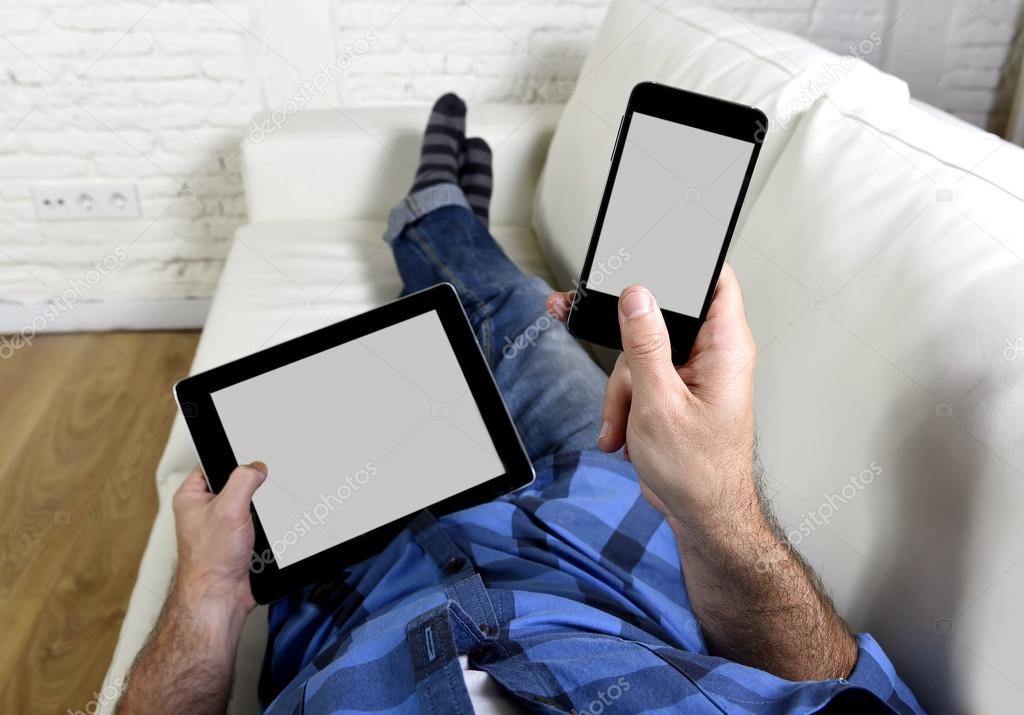 man lying on couch at home living room using simultaneously mobile phone and digital tablet pad in internet technology