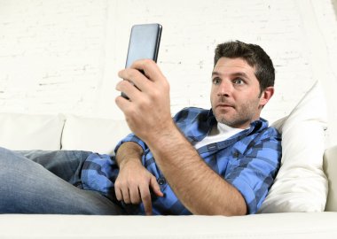 young attractive man lying relaxed at home couch texting and surfing on internet in his mobile phone