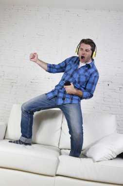 20s or 30s man jumped on couch listening to music on mobile phone with headphones playing air guitar