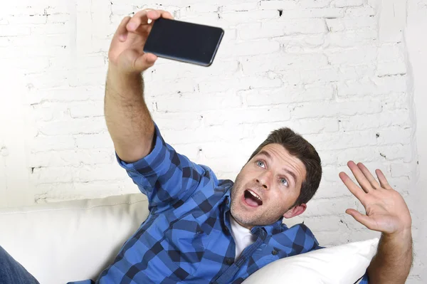 Young attractive 30s man taking selfie picture or self video with mobile phone at home sitting on couch smiling happy — Stock fotografie