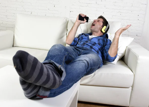 Happy crazy man on couch listening to music holding mobile phone as microphone — Stock fotografie