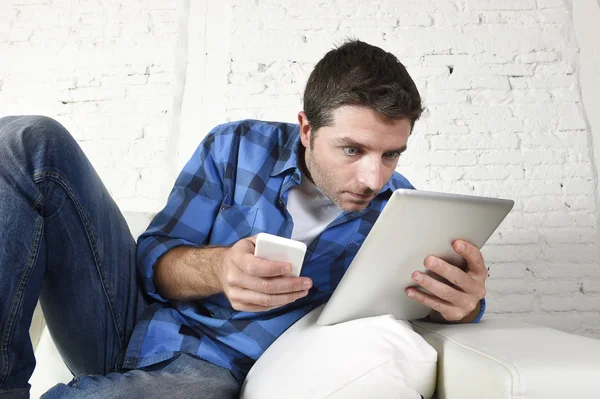 Giovane uomo a casa divano che lavora con il telefono cellulare e tablet digitale sovraccarico di stress — Foto Stock