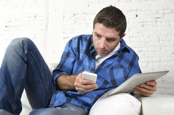 Giovane uomo a casa divano che lavora con il telefono cellulare e tablet digitale sovraccarico di stress — Foto Stock