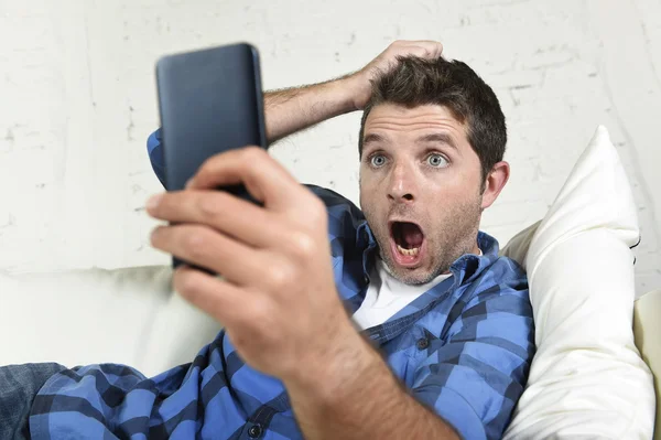 Young attractive man lying at home couch using internet on mobile phone looking surprised and shocked — Stockfoto