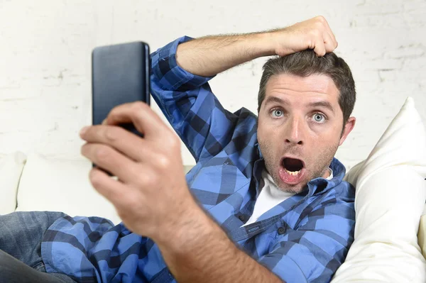 Young attractive man lying at home couch using internet on mobile phone looking surprised and shocked — Stock fotografie