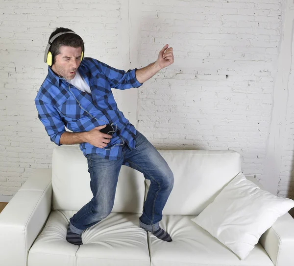 20s or 30s man jumped on couch listening to music on mobile phone with headphones playing air guitar — Stock fotografie