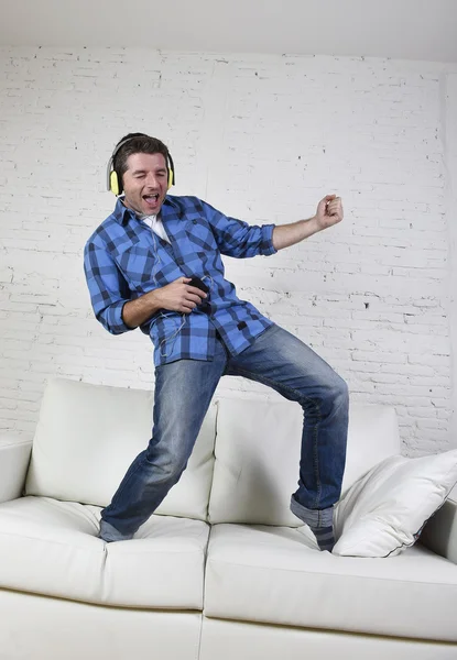 20s or 30s man jumped on couch listening to music on mobile phone with headphones playing air guitar — Stock Photo, Image