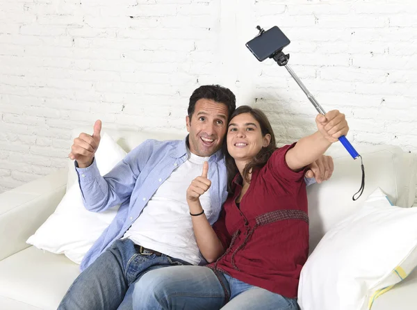 Atractiva pareja tomando foto selfie o disparando auto video con teléfono móvil y palo sentado en el sofá de casa sonriendo feliz —  Fotos de Stock