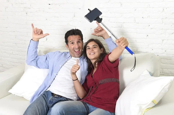 Attractive couple taking selfie photo or shooting self video with mobile phone and stick sitting at home couch smiling happy — Stockfoto