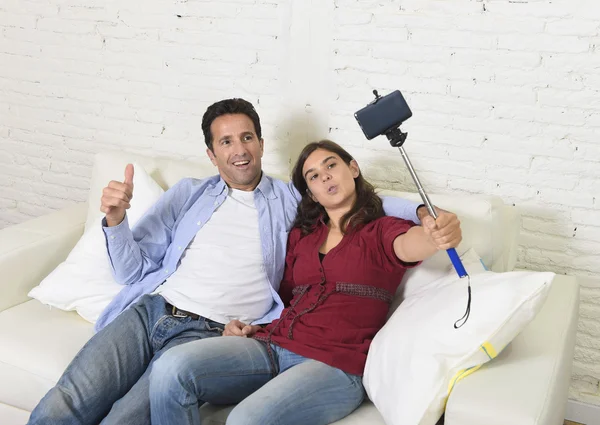 Attractive couple taking selfie photo or shooting self video with mobile phone and stick sitting at home couch smiling happy — Zdjęcie stockowe