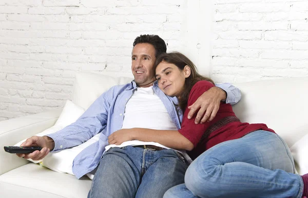 Couple in love cuddling on home couch relaxing watching movie on television with man holding remote control — Zdjęcie stockowe