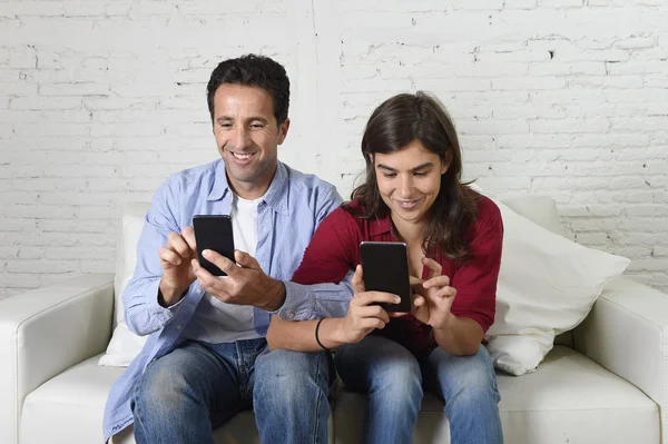 Young attractive couple of man and woman together at home couch with arms interlaced using mobile phone compulsively — Stockfoto