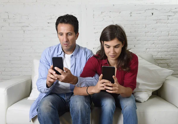 Joven pareja atractiva de hombre y mujer juntos en el sofá de casa con los brazos entrelazados usando el teléfono móvil compulsivamente —  Fotos de Stock
