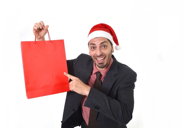 Young attractive businessman in Christmas Santa hat holding and pointing red shopping bag in December and New year sale — Stockfoto