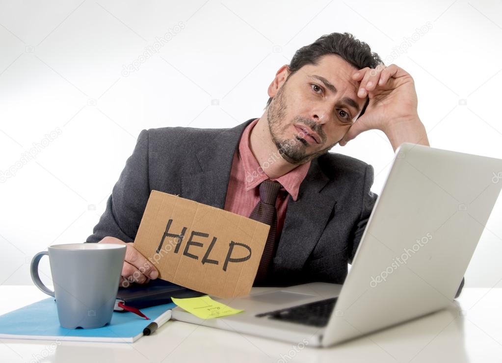 businessman in suit and tie sitting at office desk working on computer laptop asking for help holding cardboard sign