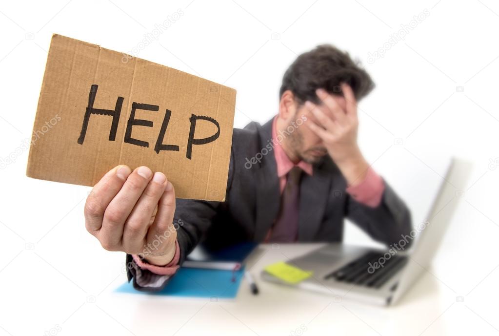 businessman in suit and tie sitting at office desk working on computer laptop asking for help holding cardboard sign