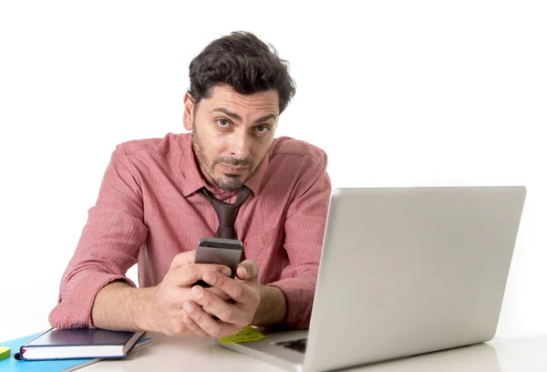 Jovem empresário atraente trabalhando na mesa de escritório com telefone celular e computador portátil olhando entediado e cansado — Fotografia de Stock