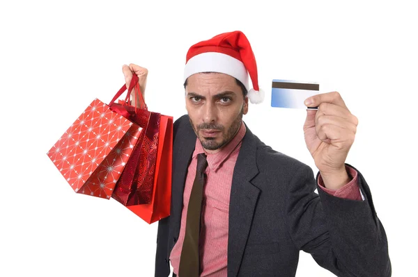 Businessman in Santa Claus Christmas hat holding shopping bags and credit card in worried face expression overwhelmed and in stress — ストック写真