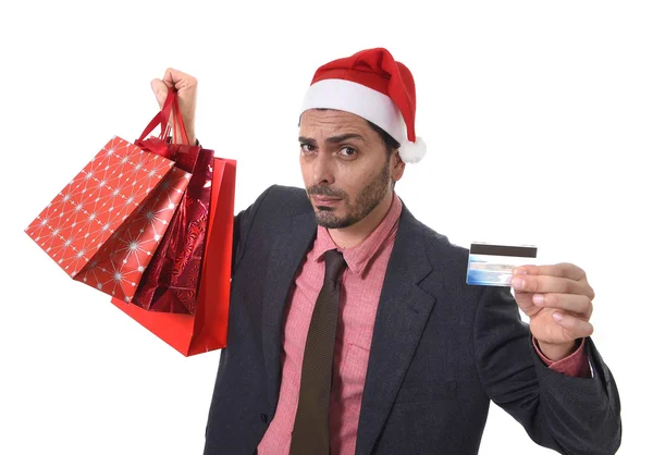 Businessman in Santa Claus Christmas hat holding shopping bags and credit card in worried face expression overwhelmed and in stress — ストック写真