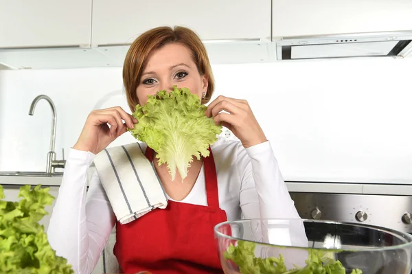 Jovem mulher bonita em avental vermelho em casa cozinha preparando tigela de salada de legumes brincando com alface — Fotografia de Stock