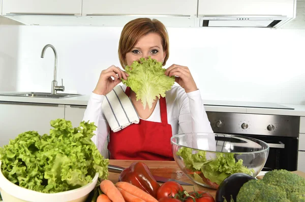 Giovane bella donna in grembiule rosso a casa cucina preparare insalata di verdure ciotola giocare con con lattuga — Foto Stock