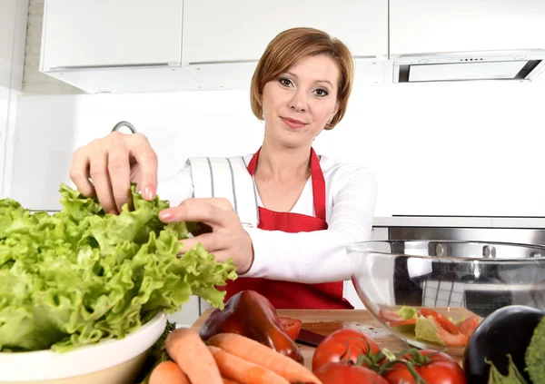 Young beautiful woman in red apron at home kitchen preparing vegetable salad bowl smiling happy — Stockfoto