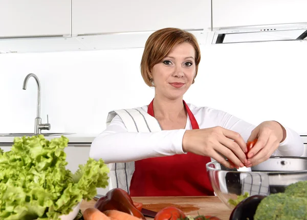 Young beautiful woman in red apron at home kitchen preparing vegetable salad bowl smiling happy — Stockfoto