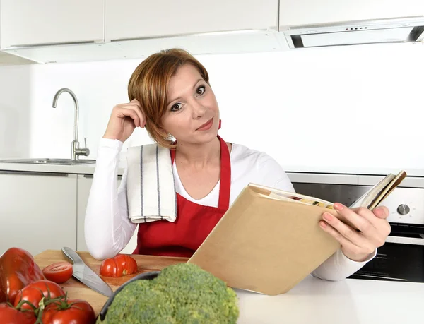 Young beautiful home cook woman in red apron at modern domestic kitchen reading cookbook following recipe — Stockfoto