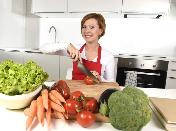 Happy woman at home kitchen preparing vegetable salad with lettuce, carrots and slicing tomato smiling — Stockfoto