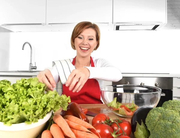 Young beautiful woman in red apron at home kitchen preparing vegetable salad bowl smiling happy — Stock Photo, Image