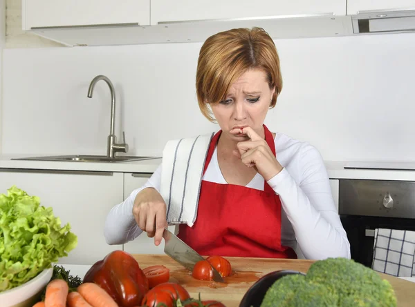 Домашня кулінарна жінка в червоному фартусі нарізає моркву з кухонним ножем, страждає від домашньої аварії різанням болючого пальця — стокове фото