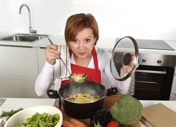Счастливый дом повар женщина в красном фартуке на домашней кухне держа кастрюлю с супом дегустации вкусное овощное рагу — стоковое фото