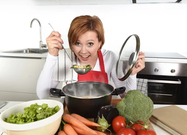 Счастливый дом повар женщина в красном фартуке на домашней кухне держа кастрюлю с супом дегустации вкусное овощное рагу — стоковое фото
