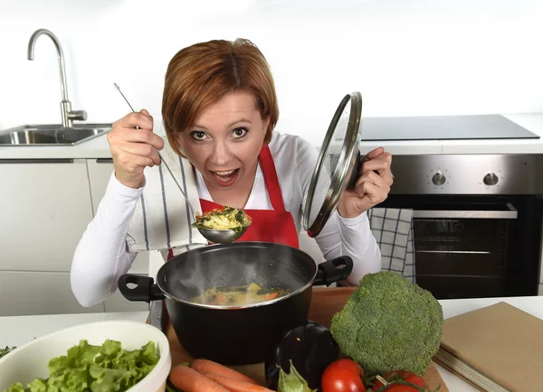 Casa feliz cozinheiro mulher em avental vermelho na cozinha doméstica segurando panela com sopa degustação delicioso guisado vegetal — Fotografia de Stock