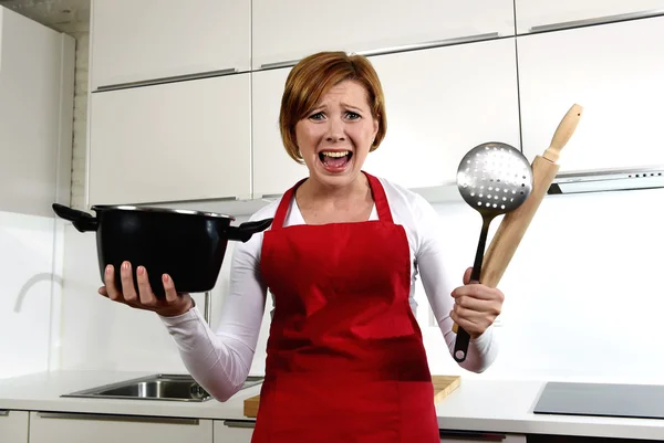 Rookie home cook woman in red apron at home kitchen holding cooking pan and rolling pin screaming desperate in stress — Stockfoto