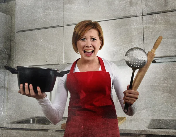 Young attractive rookie home cook woman in red apron at home kitchen holding cooking pan and rolling pin screaming desperate in stress confused and helpless in lifestyle and cooking mess — Stockfoto