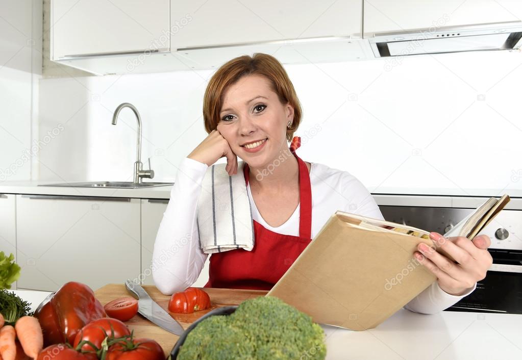 young beautiful home cook woman in red apron at modern domestic kitchen reading cookbook following recipe